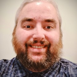 Headshot of cheerful man with very short hair, brown eyebrows, moustache and beard, smiling at the camera.